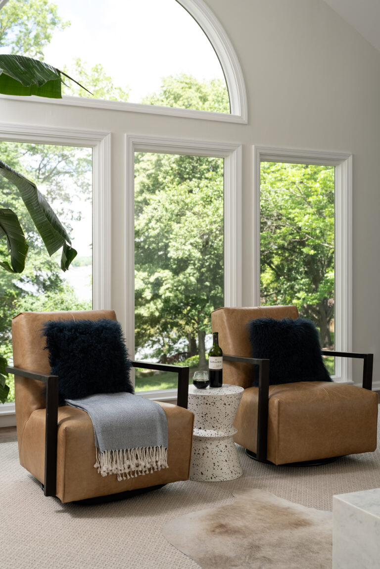 Two light caramel leather chairs are styled with textured black pillows in front of an architectural picture window.